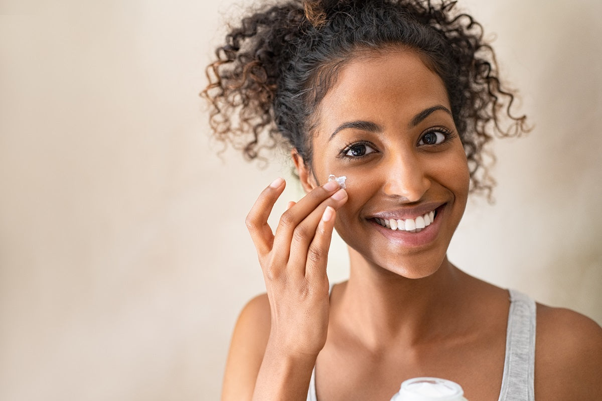 Smiling african girl with curly hair applying facial moisturizer while holding jar and looking at camer